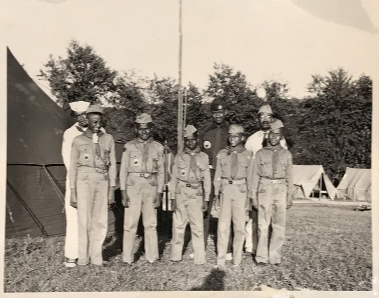 Item #66317 FIVE 12-15 YEAR OLD AFRICAN-AMERICAN BOY SCOUTS, in full uniform (khakis, side caps, kerchiefs, lanyards, official badges, etc.), along with three older African-American leaders, also in full uniform, as pictured in a large format photograph, 8 x 10 inches, standing in a campground, with tents and a stand of trees in the background. Scouting, Photograph, African-Americana.